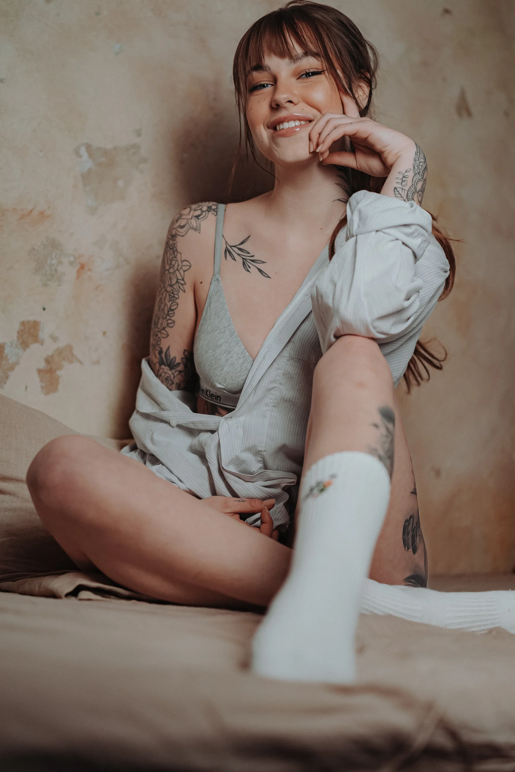 Eine tätowierte Frau sitzt entspannt auf einem Bett in Unterwäsche und einem offenen Hemd. Sie lächelt selbstbewusst und blickt in die Kamera.<br />
