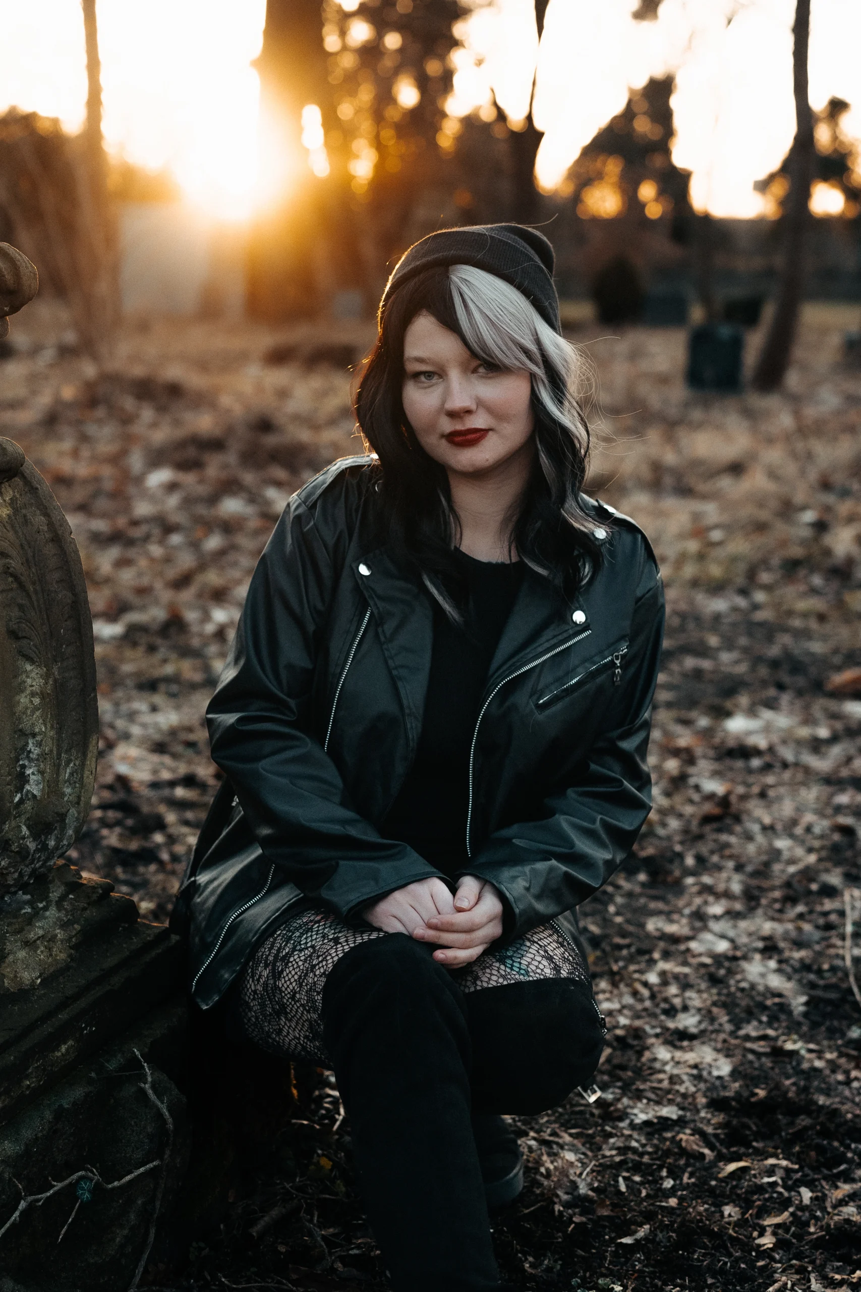 Eine Frau mit zweifarbigen Haaren, rotem Lippenstift und dunkler Kleidung sitzt im herbstlichen Wald. Die Sonne geht im Hintergrund unter und wirft warmes Licht auf die Szene.<br />
