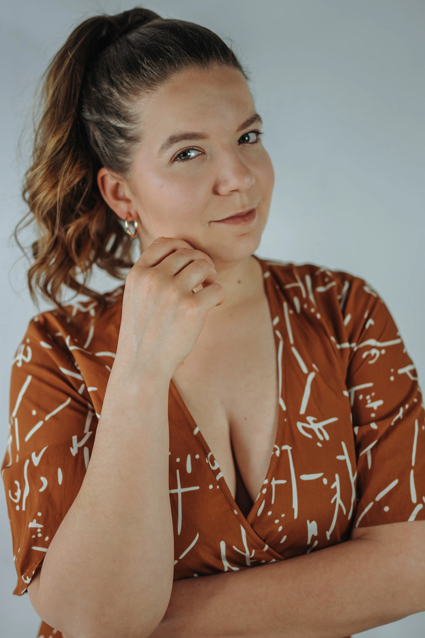 Eine Frau mit hochgebundenem Haar und braunem gemustertem Kleid blickt direkt in die Kamera. Sie hält nachdenklich eine Hand am Kinn und wirkt selbstbewusst.
