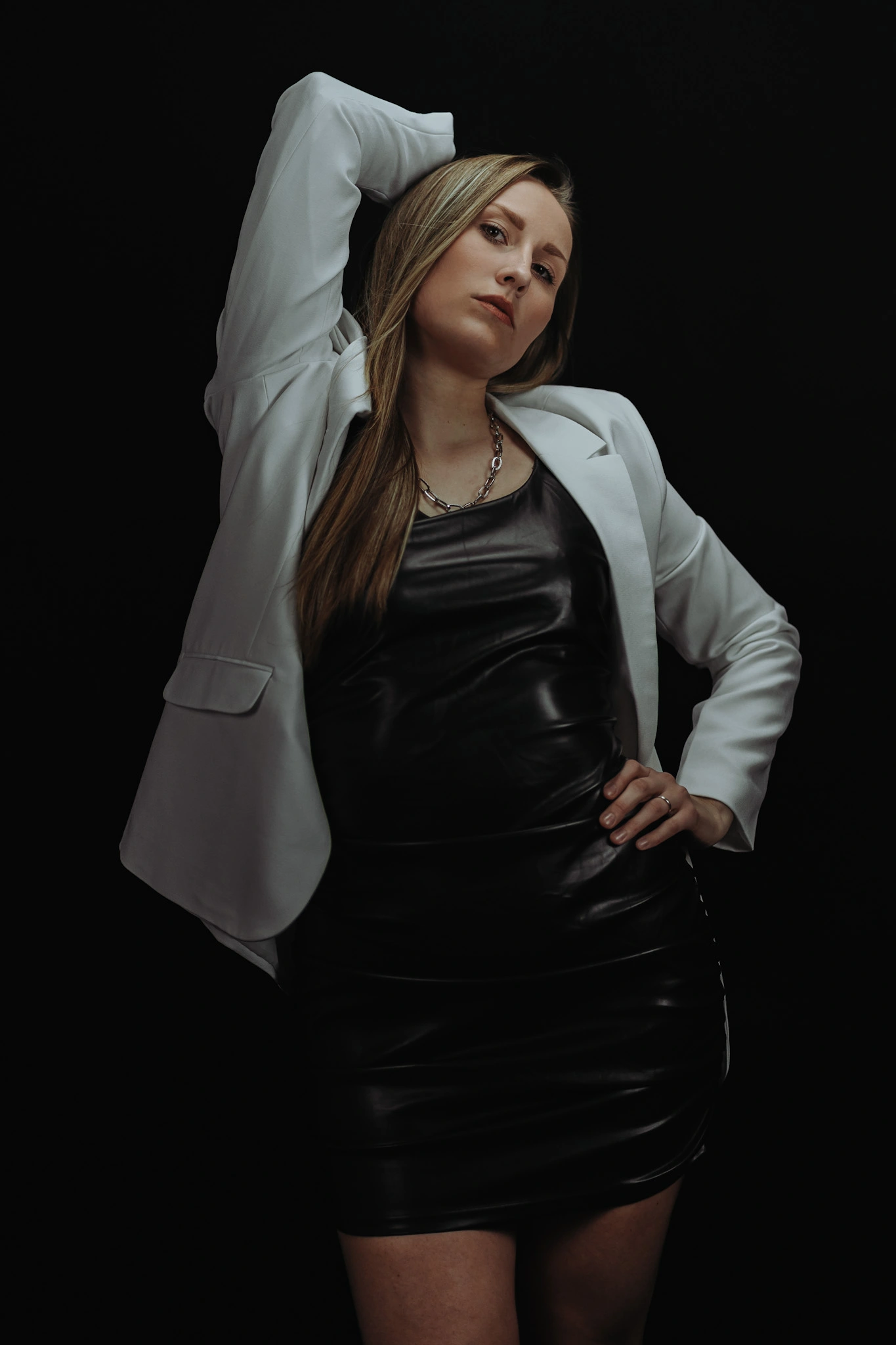 Eine Frau in schwarzem Lederkleid und weißem Blazer posiert selbstbewusst vor einem schwarzen Hintergrund. Sie blickt ernst und kraftvoll in die Kamera.<br />
