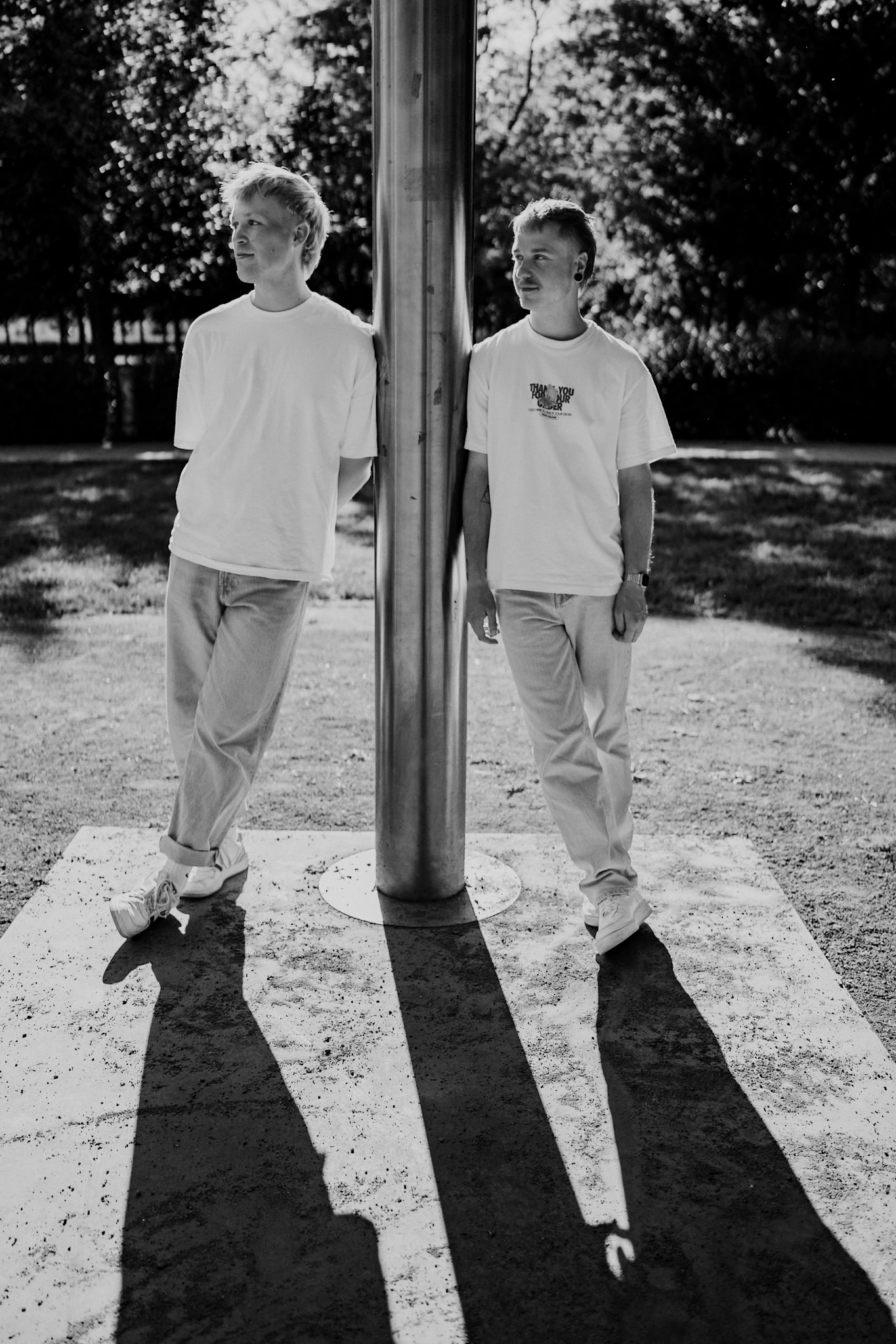 Zwei junge Männer in weißen T-Shirts und Jeans stehen entspannt an einem Metallpfeiler im Freien. Die Szene ist in Schwarz-Weiß aufgenommen, das Licht wirft lange Schatten.<br />
