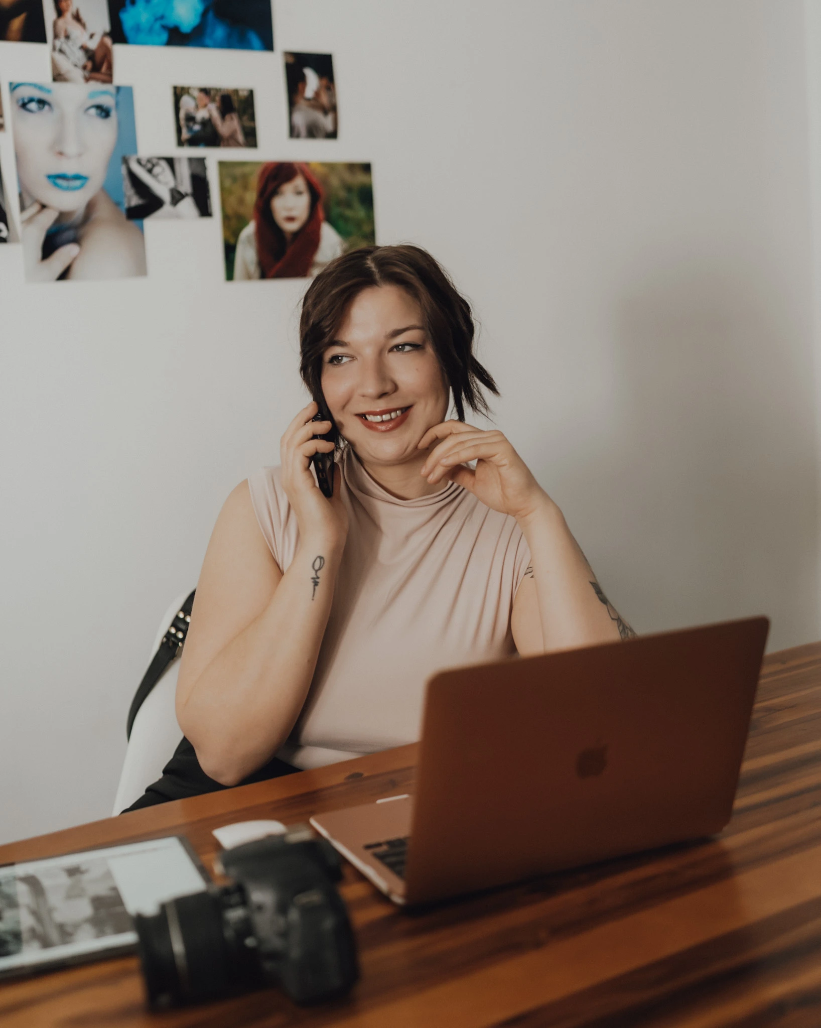Eine Fotografin sitzt an einem Schreibtisch mit Laptop, Kamera und Smartphone. Sie telefoniert lächelnd und wirkt entspannt und professionell.