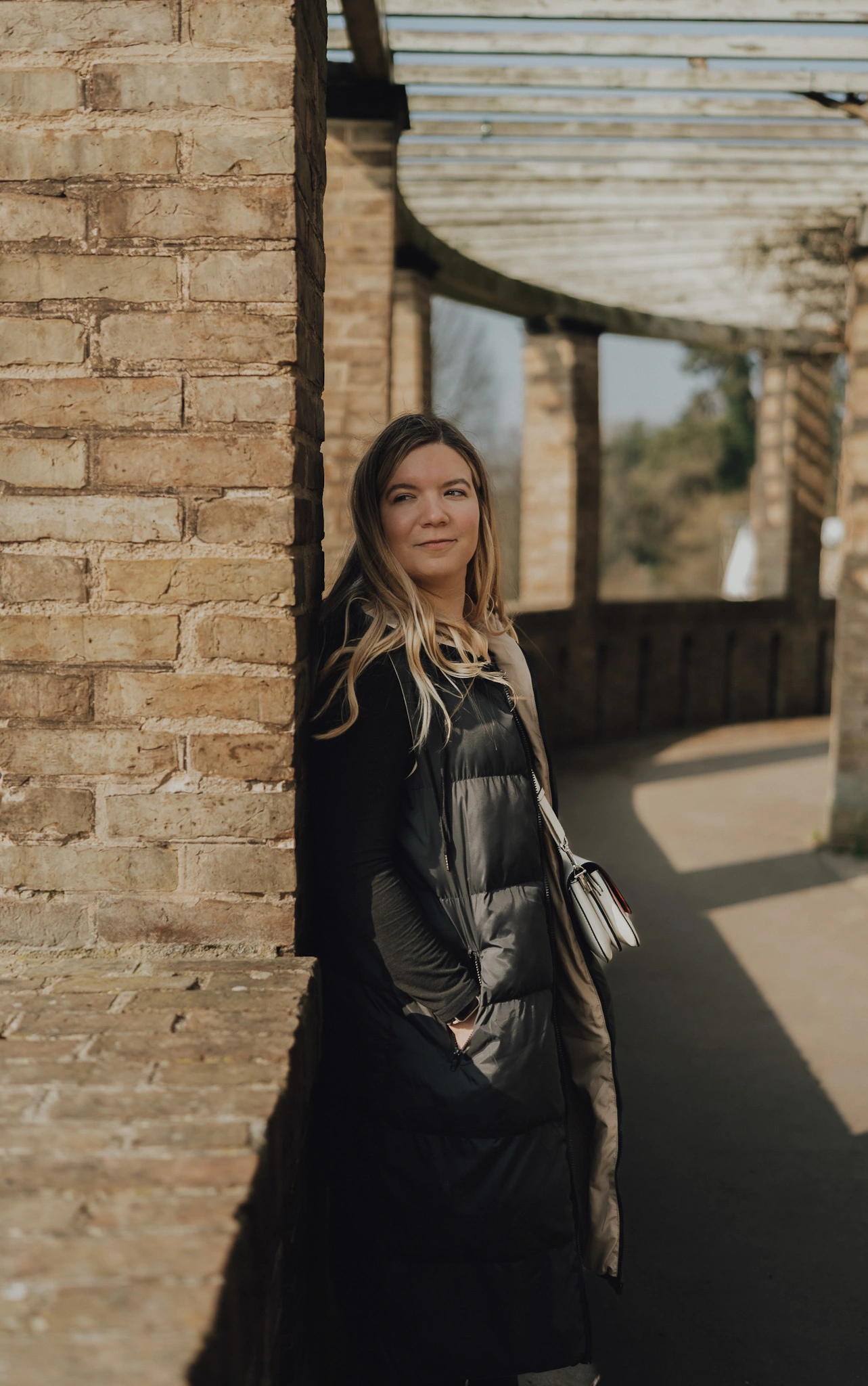 Eine Frau mit langen blonden Haaren lehnt entspannt an einer Backsteinwand in einem überdachten Säulengang. Sonnenlicht wirft klare Schatten auf den Boden.