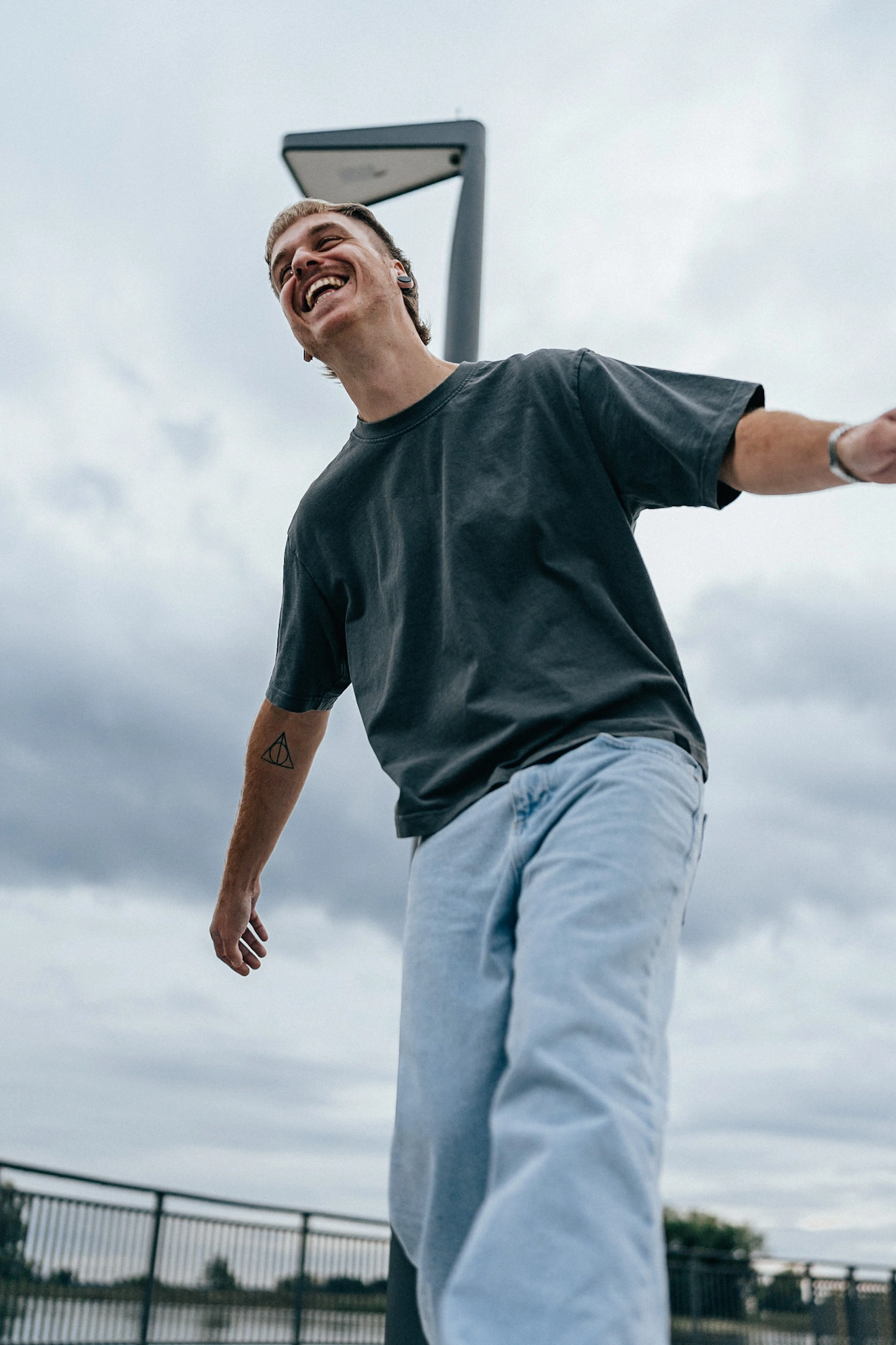 Junger Mann lacht freiherzig mit ausgebreiteten Armen im Freien unter bewölktem Himmel, im lässigen Look mit dunklem Shirt und hellblauer Jeans.