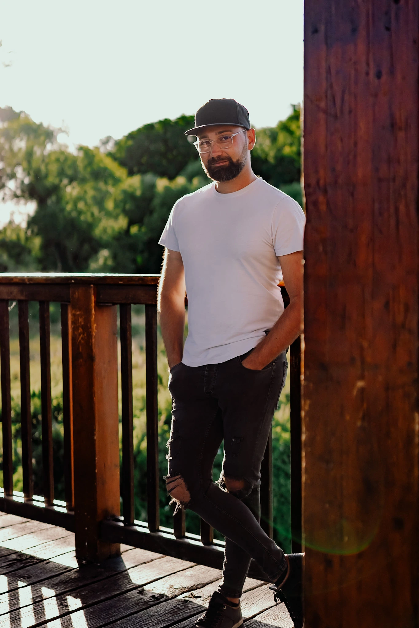 Mann mit Brille und Cap lehnt lässig an einem Holzpfosten auf einer Veranda im Gegenlicht der Abendsonne.<br />
