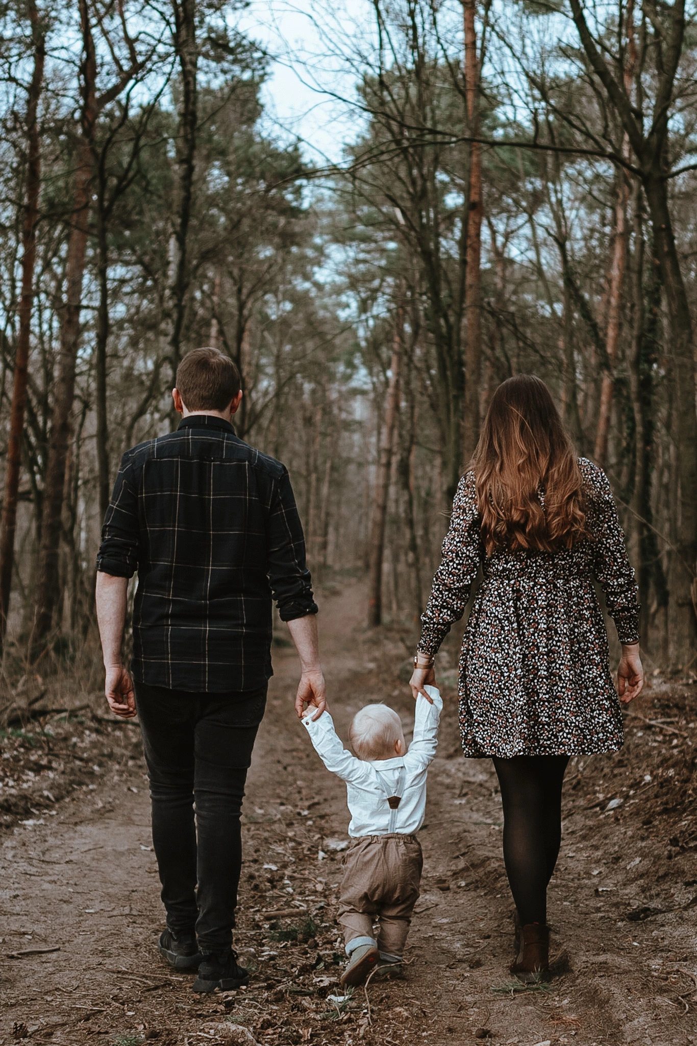 Ein Paar läuft mit einem Kleinkind, das sie an beiden Händen halten, einen Waldweg entlang. Alle sind von hinten zu sehen und gehen gemeinsam in Richtung Lichtung.<br />
