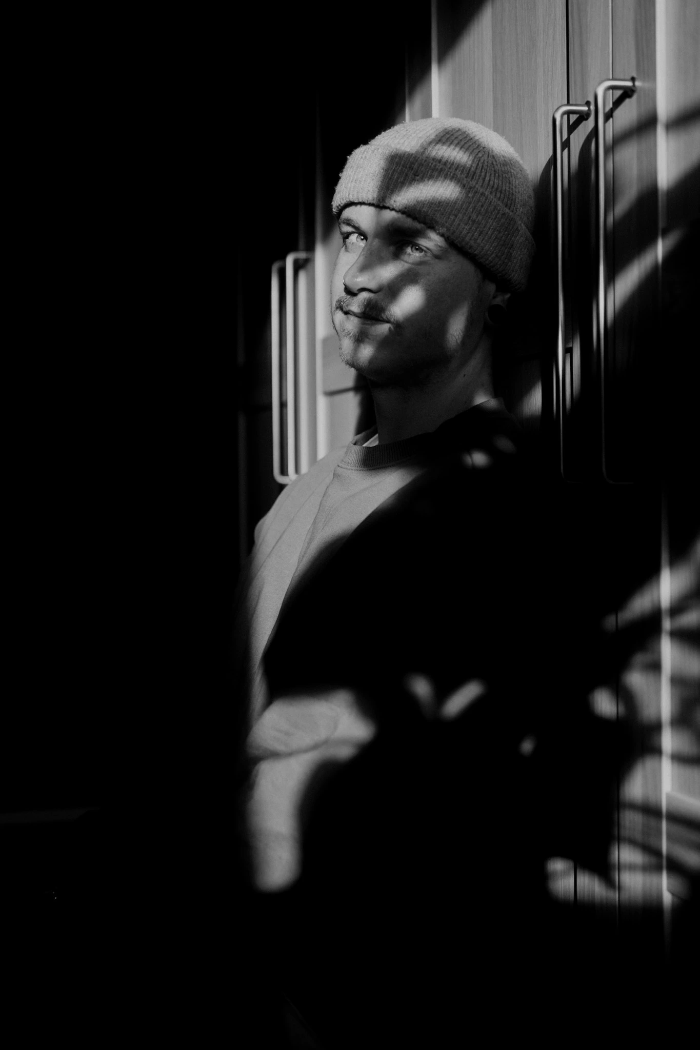 Ein Mann mit Mütze steht im Schatten an einem Schrank, während Licht durch ein Fenster Muster auf sein Gesicht wirft. Das Foto ist in Schwarz-Weiß gehalten.<br />
