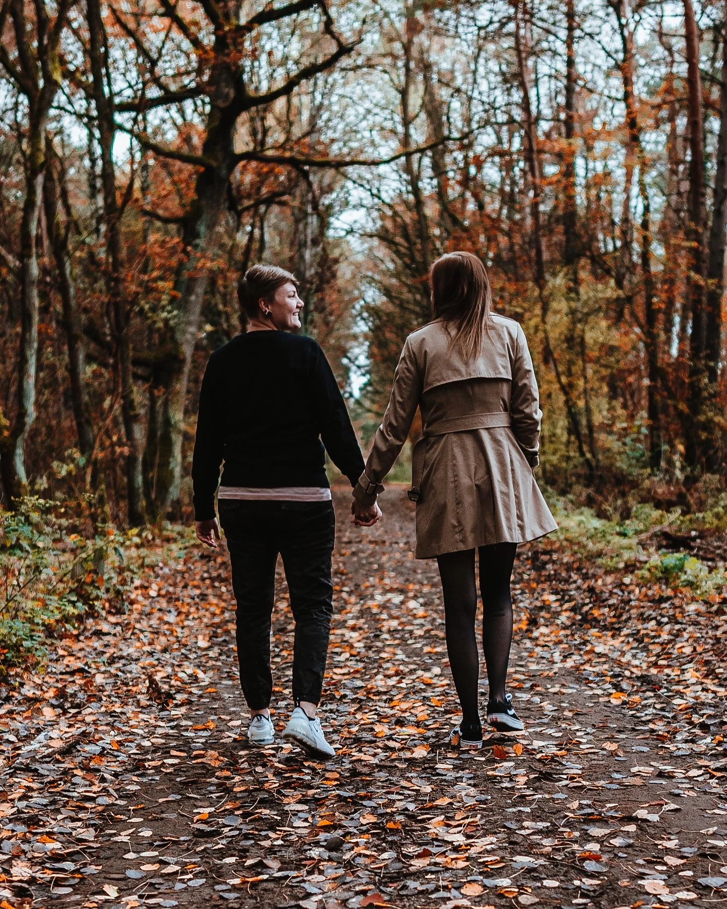 Zwei Personen gehen Hand in Hand auf einem mit Laub bedeckten Waldweg, von hinten fotografiert, inmitten herbstlich gefärbter Bäume.
