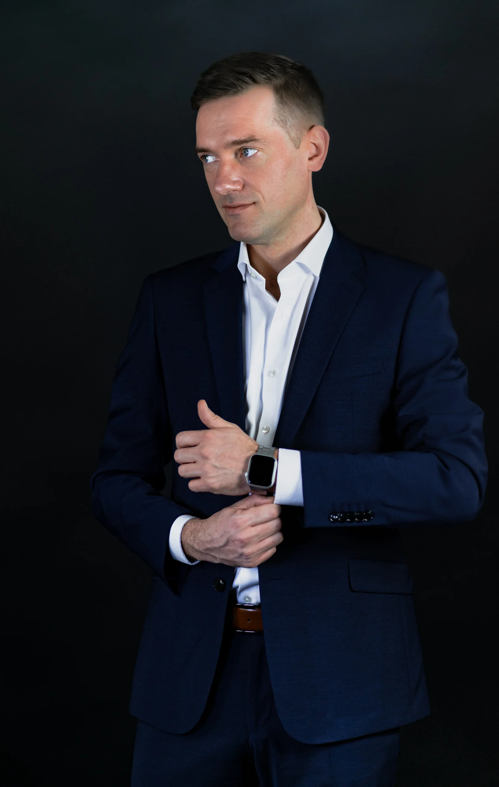 Mann in dunkelblauem Anzug mit weißem Hemd blickt zur Seite und richtet seine Smartwatch.