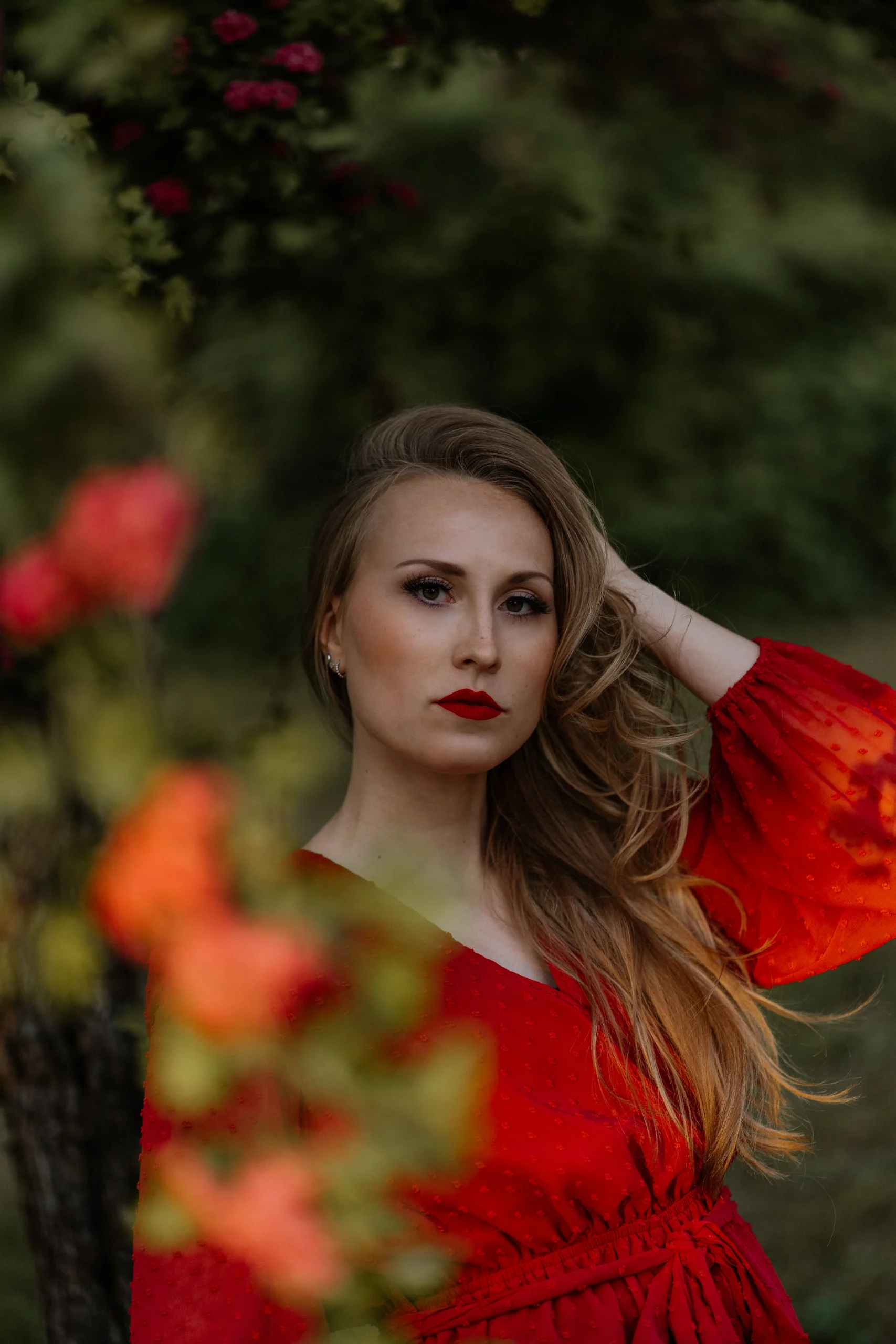 Porträt einer Frau in rotem Kleid mit offenen Haaren, stehend im Grünen, umrahmt von roten Blüten.