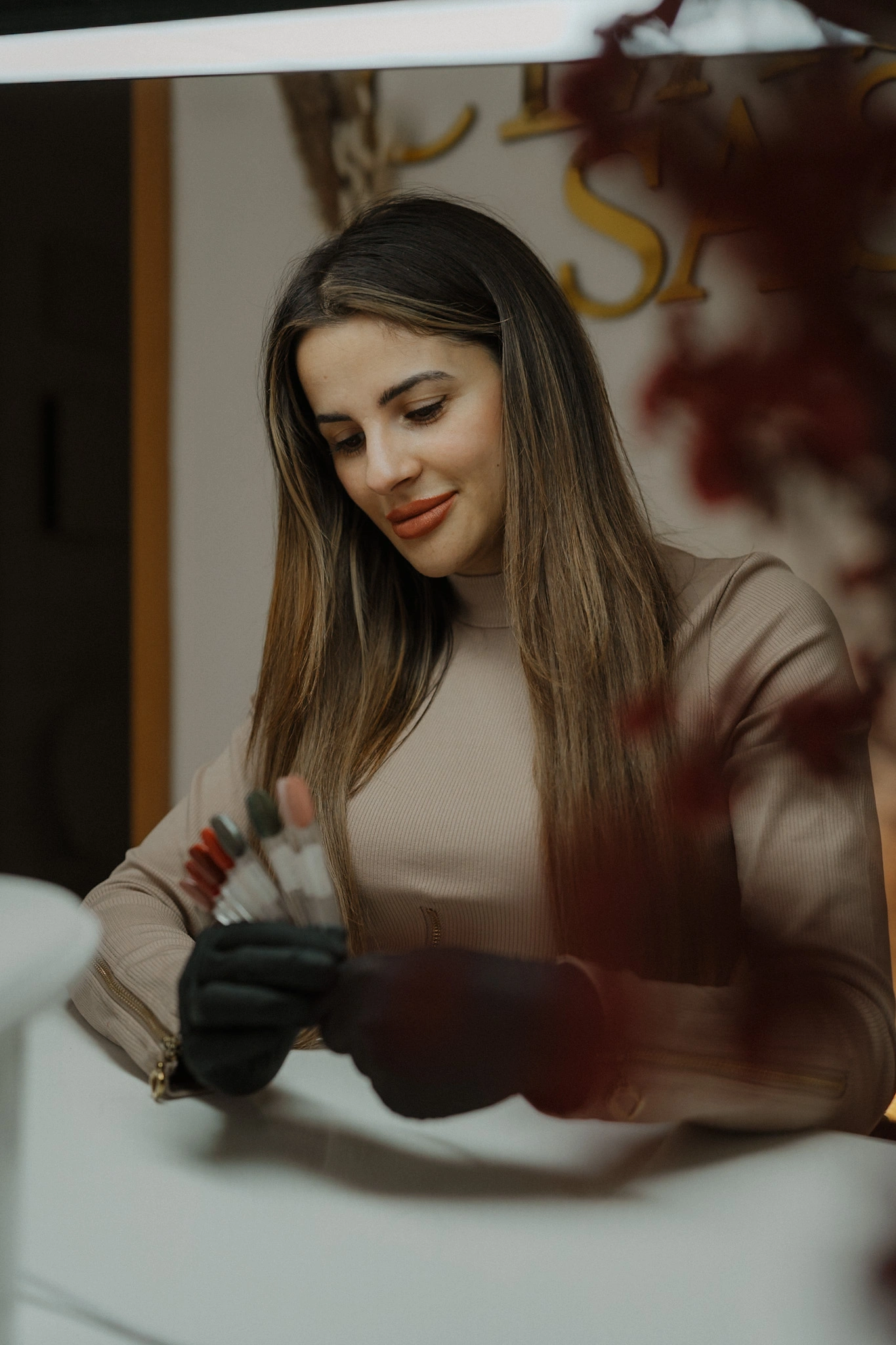 Frau mit langen Haaren und beigem Oberteil wählt Nagelfarben aus, trägt schwarze Handschuhe und sitzt an einem Nageldesign-Tisch im Studio