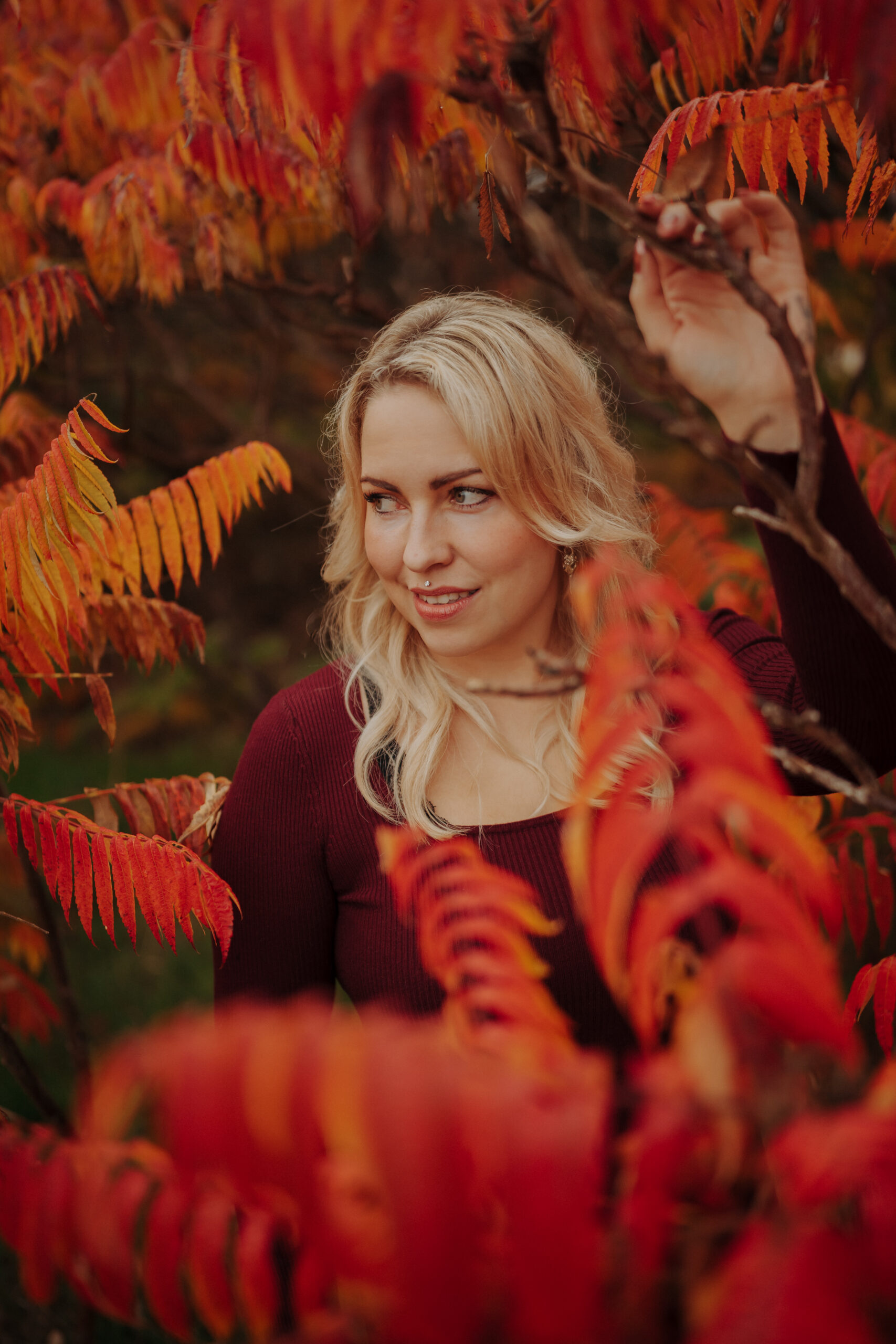Porträt einer Frau mit blondem Haar zwischen leuchtend roten und orangefarbenen Herbstblättern, stimmungsvolle Outdoor Aufnahme im warmen Herbstlicht